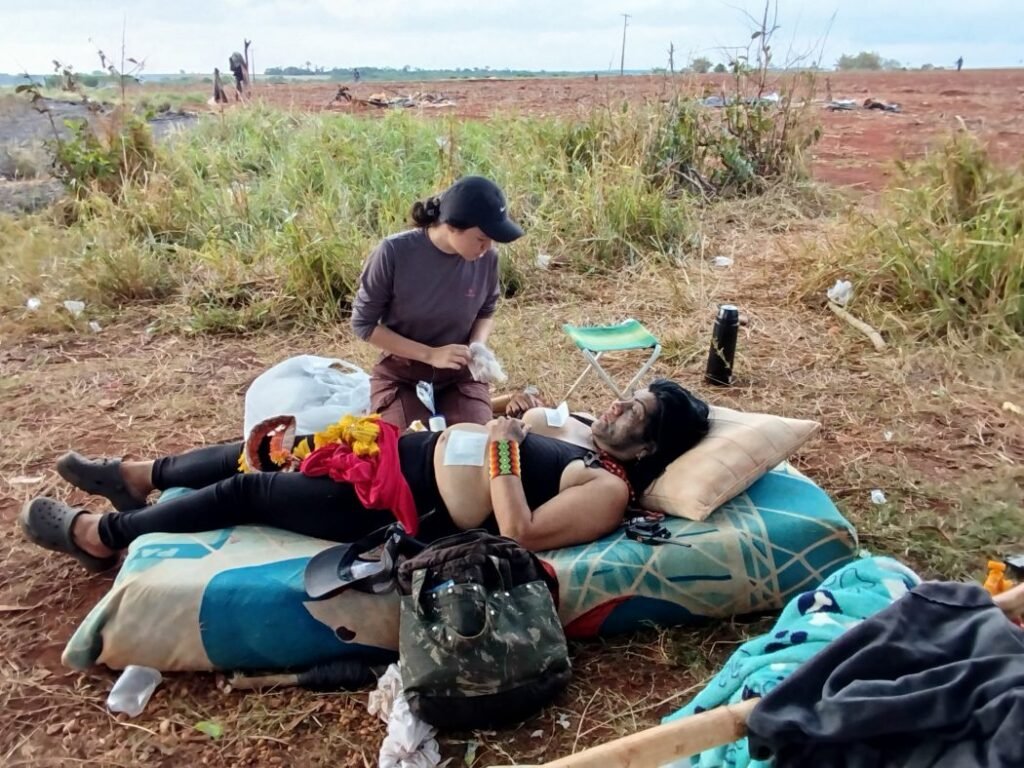 Indígena ferida na retomada de após ação da Polícia Militar, na retomada de Guyraroká. Ao fundo, os barracos destruídos (Foto: Lídia Farias/Cimi)